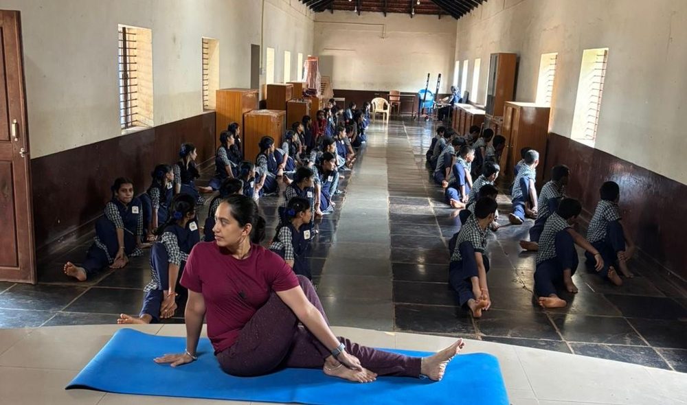 A yoga teacher demonstrating a seated twist while school children practise on the floor of a large hall