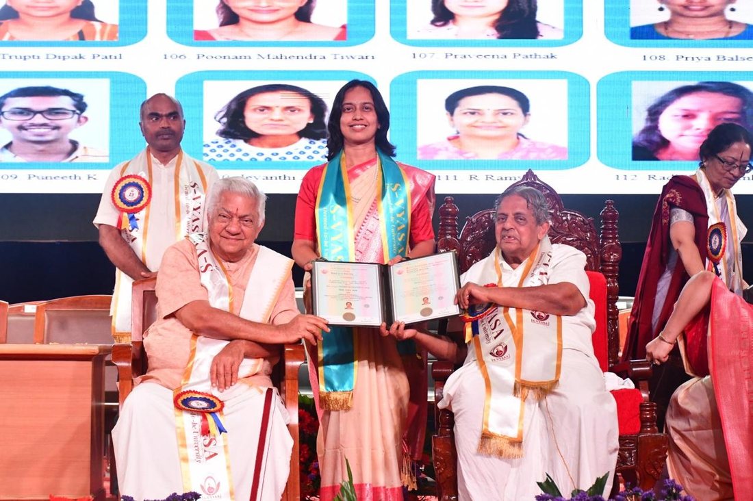 Priya Balse in an S-VYASA sash holding her degree certificate on stage at the convocation, with senior university figures seated beside her